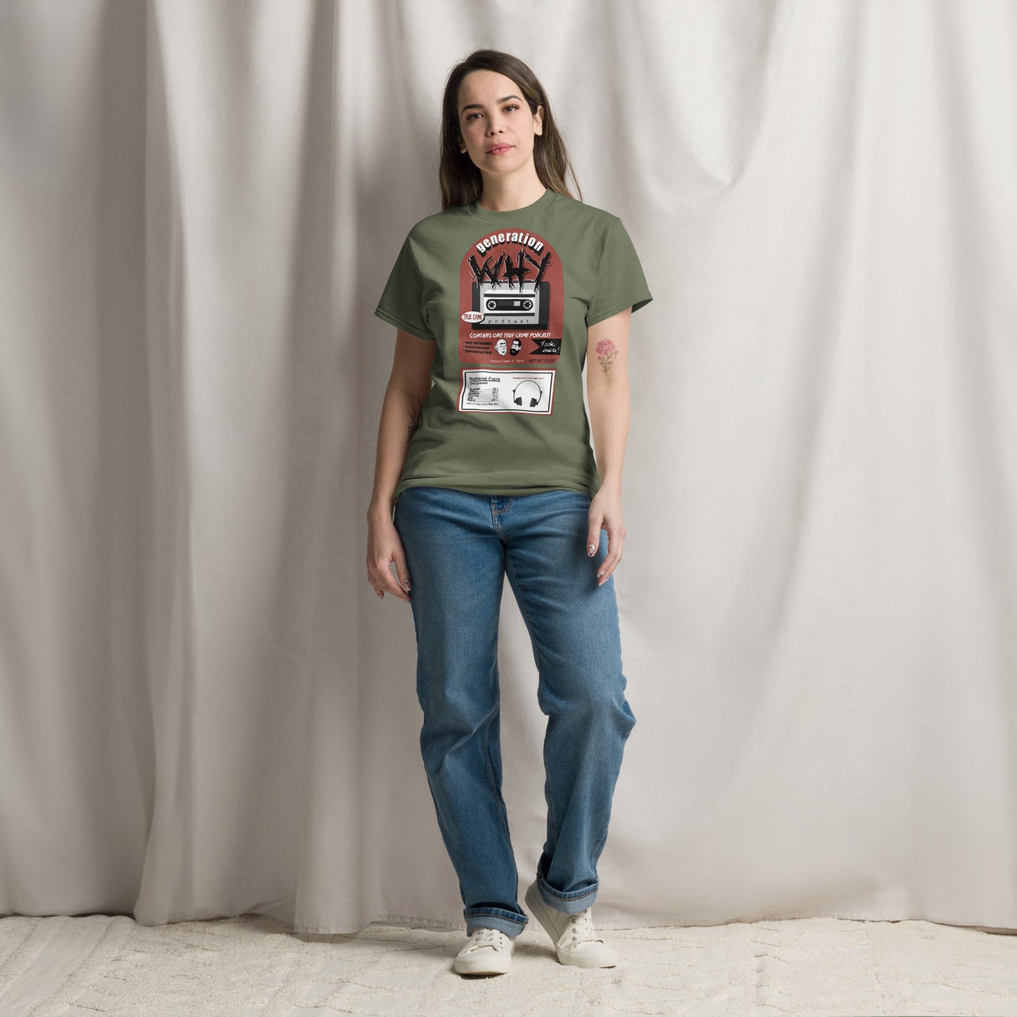 A woman stands against a draped beige fabric backdrop wearing an olive green graphic T-shirt with a cassette player print, blue jeans, and white sneakers.