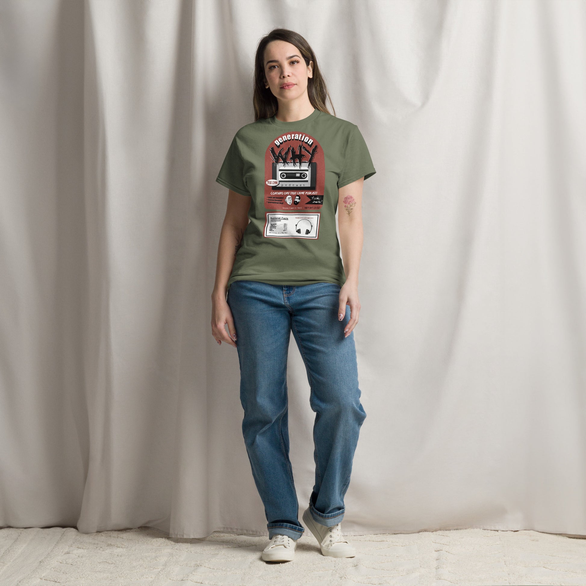 A woman stands against a draped beige fabric backdrop wearing an olive green graphic T-shirt with a cassette player print, blue jeans, and white sneakers.